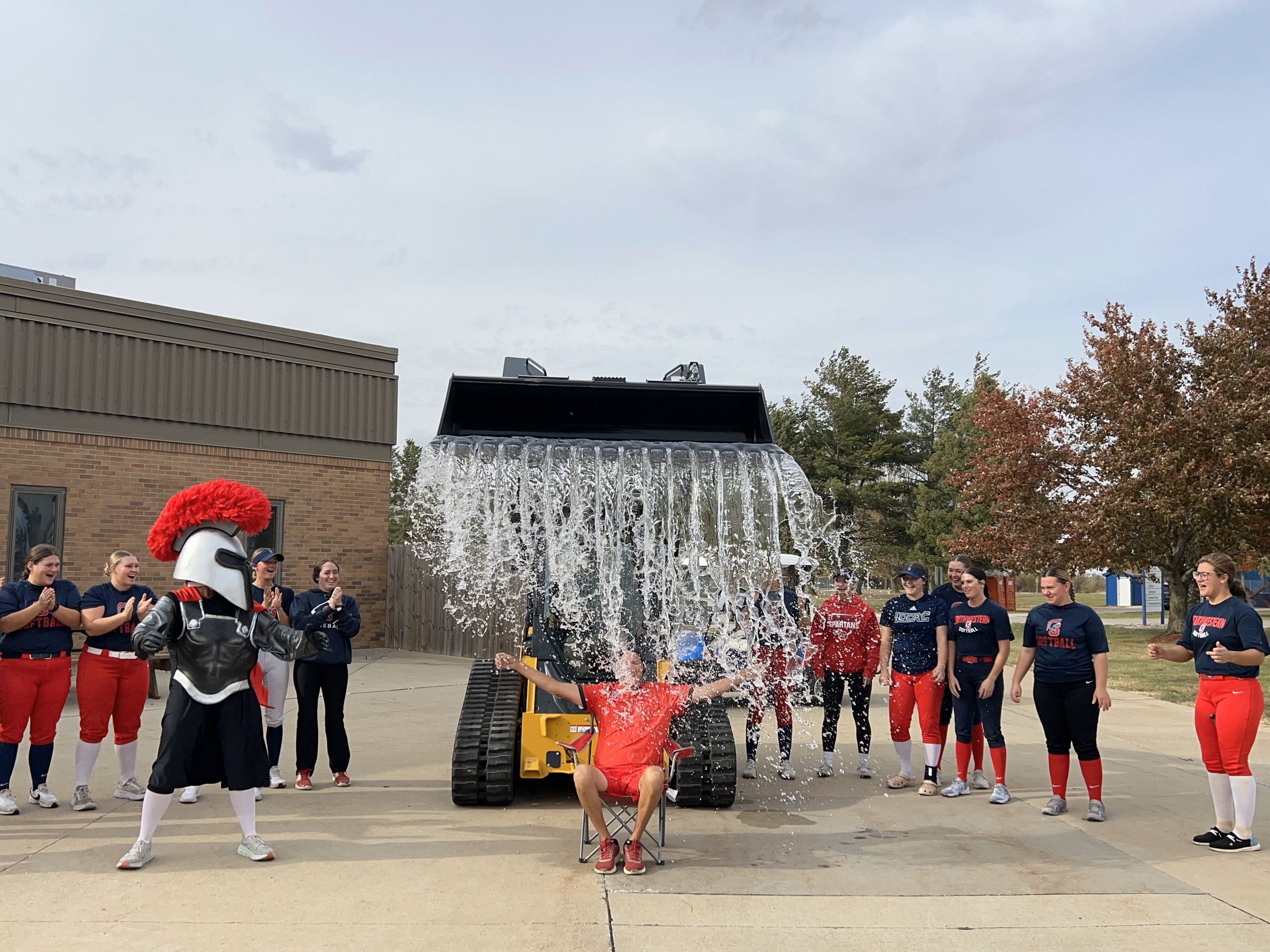 Coach Jensen sitting in a chair getting doused by a wave of water being poured from a loader over his head with the team watching